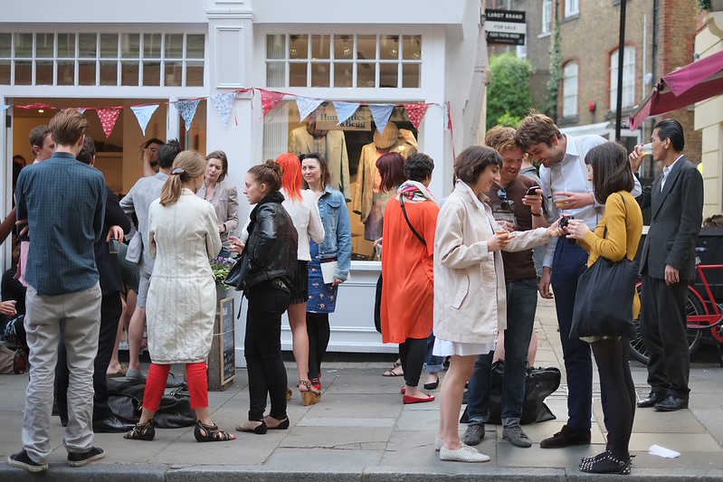 A crowd in conversation outside a London shopfront. Real people, mid-moment, available light.